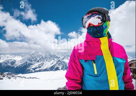 Portrait d'une fille skieuse dans une veste colorée et lumineuse dans un masque de ski avec un visage couvert par un jour ensoleillé sur le fond de Banque D'Images