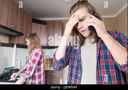 Perturbation grave jeune homme aux cheveux longs debout dans la cuisine utilise son téléphone portable ou commander un service sur le tal Internet Banque D'Images