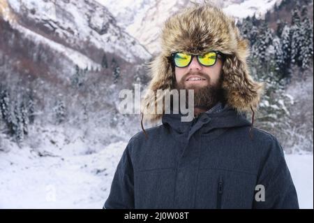 Portrait d'un jeune barbu attrayant dans un grand chapeau de fourrure et des lunettes de soleil contre la forêt enneigée dans les montagnes Banque D'Images