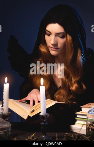 Moyenne gros plan attrayante caucasienne fille avec de longs cheveux dorés dans une cagoule noire est assis à une table et des feuilles à travers un livre. Costume W Banque D'Images