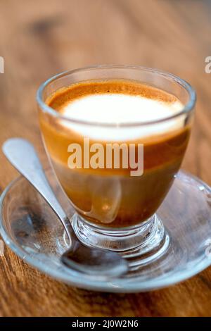 Boisson au café macchiato avec mousse et couches dans une tasse en verre avec une cuillère sur fond de bois. Cryptographie macro, mise au point sélective, gros plan et flou avec bokeh Banque D'Images