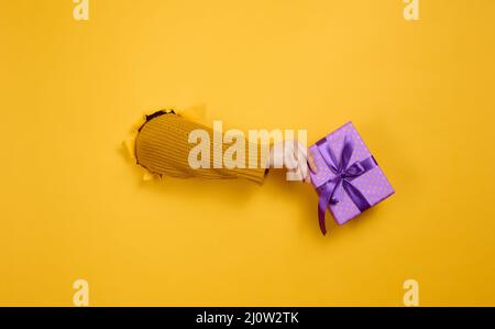 La main femelle tient une boîte avec un cadeau sur fond jaune, une partie du corps dépasse d'un trou déchiré dans un fond en papier. Co Banque D'Images