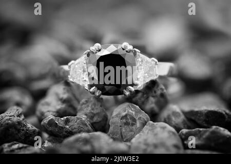 Un anneau de diamant noir avec des diamants blancs de mille milliards de coupe reposant sur une pile de pierres précieuses brutes. Image monotone. Banque D'Images