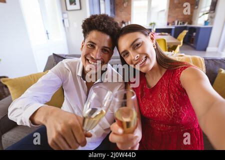 Portrait d'un jeune couple biracial souriant avec flûtes à champagne emportant le selfie à la maison pendant la fête Banque D'Images