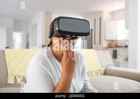 Femme afro-américaine âgée utilisant le simulateur de réalité virtuelle avec la main sur le menton à la maison Banque D'Images