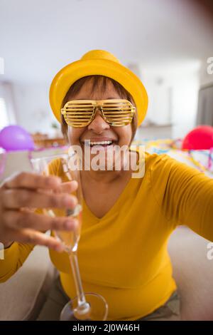 Bonne femme senior dans des lunettes de protection en train de passer un appel vidéo tout en dégustant du champagne pendant la fête à la maison Banque D'Images