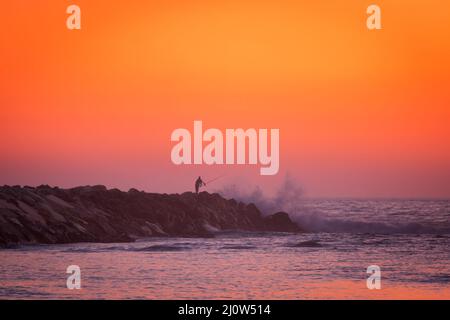 Pêche de silhouette de pêcheur dans l'océan atlantique avec des vagues s'écrasant sur un quai en pierre au coucher du soleil, au Portugal Banque D'Images