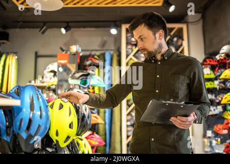 Un directeur de boutique de vélos de sexe masculin effectue un inventaire des casques de sport dans une boutique de vélos. Le propriétaire d'un magasin de sport avec un presse-papiers en hi Banque D'Images