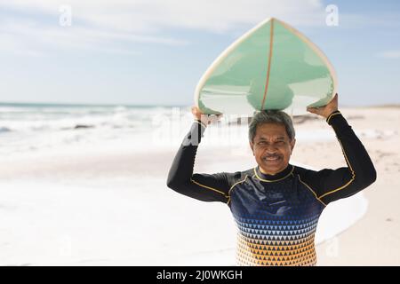 Portrait d'une femme biraciale âgée souriante portant une combinaison transportant une planche de surf sur la tête à la plage ensoleillée Banque D'Images