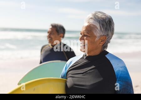 Femme aîée souriante d'oiseaux portant une planche de surf avec un homme à la plage qui regarde loin par beau temps Banque D'Images