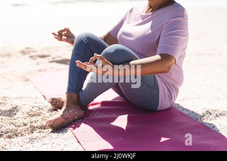Basse section de la femme biraciale senior méditant en position de lotus à la plage le jour ensoleillé Banque D'Images