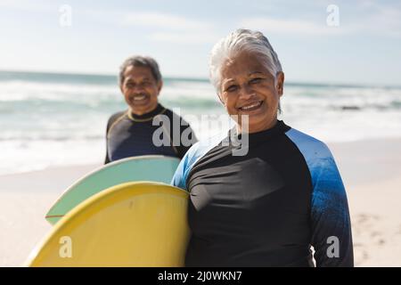 Portrait d'une femme et d'un homme biracial souriant portant des planches de surf à la plage par beau temps Banque D'Images