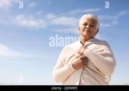 Vue à angle bas de la femme biraciale senior portant un haussement d'épaules, en regardant la plage contre le ciel bleu Banque D'Images