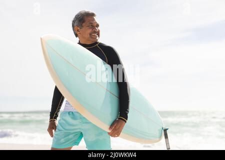 Homme biracial senior souriant portant une planche de surf et regardant la plage contre le ciel pendant la journée ensoleillée Banque D'Images