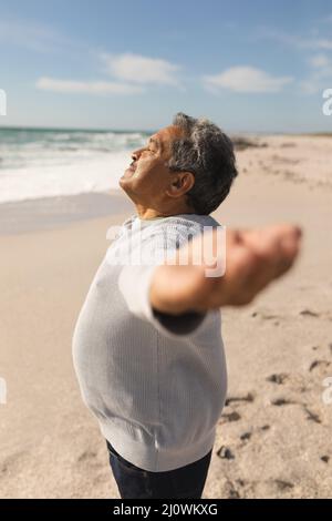 Vue latérale de l'homme aîné biracial avec les yeux fermés et les bras s'étirent à la plage le jour ensoleillé Banque D'Images