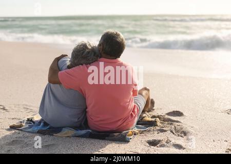 Vue arrière de l'homme biracial senior assis avec le bras autour de la femme profitant de la retraite à la plage Banque D'Images