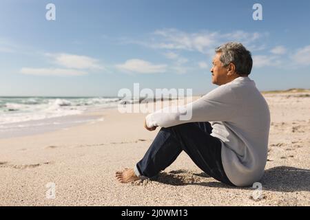 Vue latérale complète de l'homme aîné biracial assis sur le sable à la plage pendant la journée ensoleillée Banque D'Images