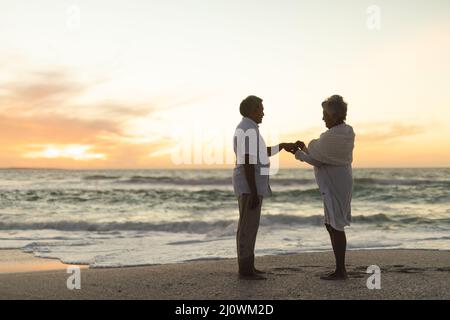 Vue latérale de la mariée biraciale senior mettant l'anneau de mariage sur le doigt du marié à la plage pendant le coucher du soleil Banque D'Images
