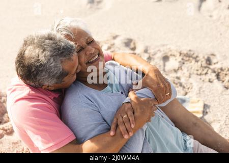 Romantique biracial homme senior embrassant la femme heureuse senior de derrière tout en s'asseyant à la plage Banque D'Images