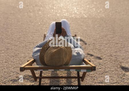 Vue en grand angle de la femme biraciale senior utilisant un smartphone tout en étant assise sur une chaise pliante à la plage Banque D'Images