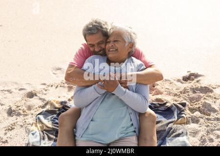 Vue en grand angle de l'aimant biracial homme senior embrassant la femme heureuse de derrière assis à la plage ensoleillée Banque D'Images