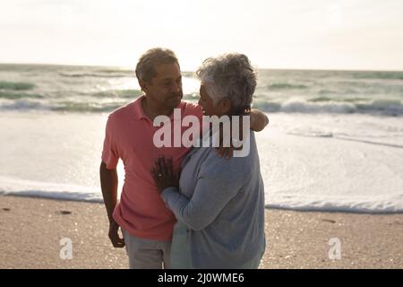 Homme biracial senior aimant regardant une femme avec le bras autour de son épaule à la plage pendant le coucher du soleil Banque D'Images