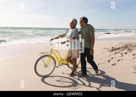 Biracial homme senior marchant et parlant avec une femme en train de rouler à vélo à la plage ensoleillée contre le ciel Banque D'Images