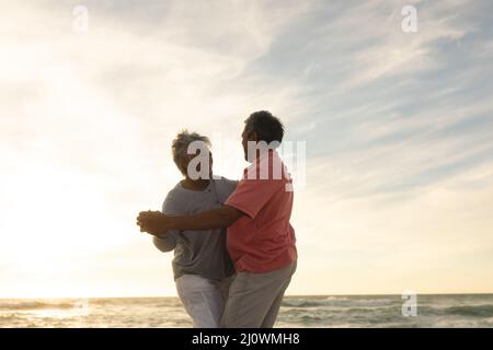 Couple biracial senior heureux dansant romantique à la plage contre le ciel pendant le coucher du soleil Banque D'Images