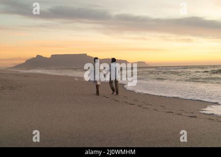 Vue arrière sur toute la longueur de couple biracial senior marchant sur la rive tout en appréciant le coucher du soleil sur la plage Banque D'Images