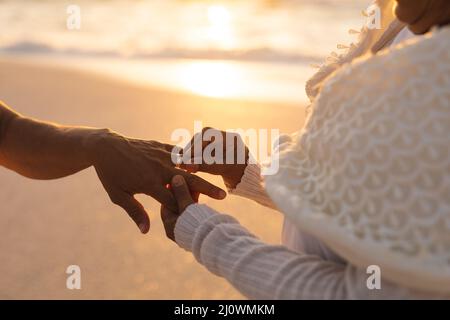 Section médiane de la mariée biraciale senior mettant l'anneau sur le doigt de l'époux à la cérémonie de mariage à la plage Banque D'Images
