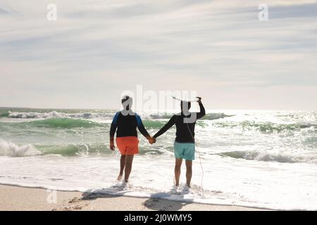 Vue arrière de l'homme biracial senior portant une planche de surf sur la tête tenant la main de la femme marchant à la plage Banque D'Images