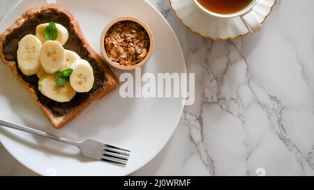 Pain de banane au chocolat avec des graines sur une assiette blanche servi avec du thé chaud sur fond de table en marbre. Banque D'Images