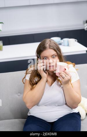 Une jeune fille en surpoids, effrayée ou stressée, parle au téléphone en tenant une tasse rose pressée sur son visage. Grosse fille parlant sur le phon Banque D'Images