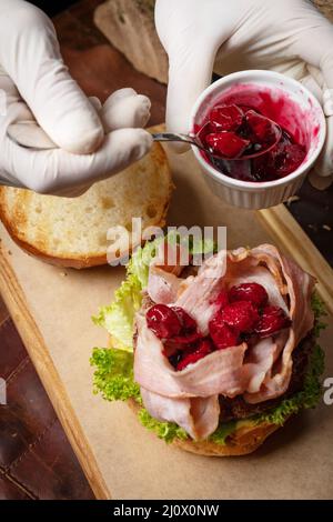 Vue de dessus.Le chef sert un hamburger de bœuf artisanal frais avec des tranches de bacon frites, recouvert de sauce aigre-douce aux cerises et de jambon frais Banque D'Images