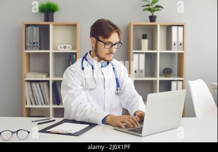 Médecin assis à table et travaillant avec des dossiers médicaux électroniques sur un ordinateur portable Banque D'Images