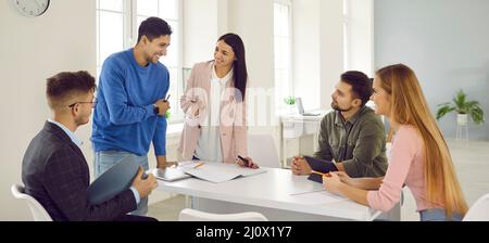 Jeunes travailleurs souriants assis au bureau, parlant et discutant des idées lors d'un briefing sur le lieu de travail. Banque D'Images