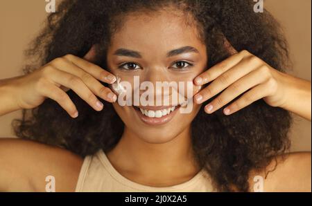 Gros plan d'une femme à la peau sombre et souriante, avec des cheveux freux qui appliquent une crème pour le visage Banque D'Images