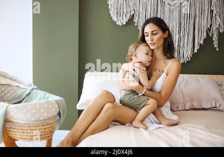 Jeune femme mère exprimant l'amour au petit bébé, assise sur le lit avec l'enfant endormi dans les mains et l'embrassant dans le front. Banque D'Images