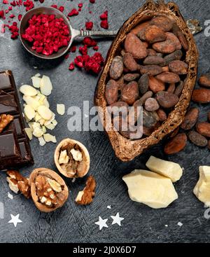 Barre de chocolat, morceaux de chocolat noir écrasés, fèves de cacao, beurre de cacao, noix, vue de dessus sur fond sombre. Banque D'Images