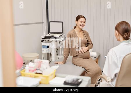 Portrait de la jeune femme enceinte souriant avec joie tout en écoutant le médecin dans la salle d'examen échographique Banque D'Images