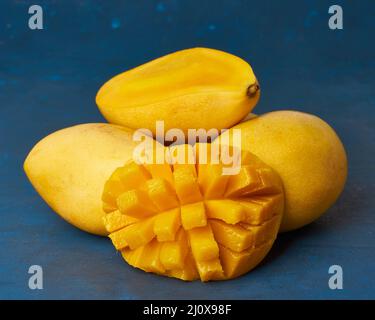 Quatre fruits de mangue entiers sur une table bleu foncé et coupés en tranches. Gros fruits jaunes mûrs juteux Banque D'Images