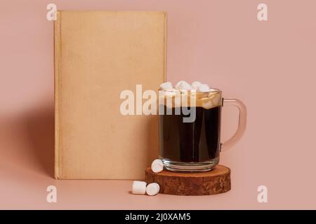 Une tasse de café en verre avec guimauves se tient sur un support en bois à côté du livre Banque D'Images