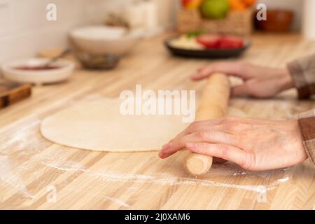 Une femme sans visage fait rouler la pâte avec une broche sur la table de cuisine à la maison, Banque D'Images