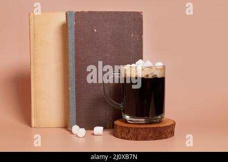 Une tasse de café en verre avec guimauves se tient sur un support en bois à côté des livres Banque D'Images