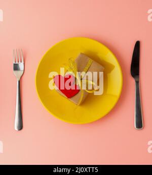 Cadeau, paquet de papier Kraft brun avec un ruban or, deux coeurs en feutre sur une assiette jaune, couteau, fourchette sur fond rose vif, Banque D'Images