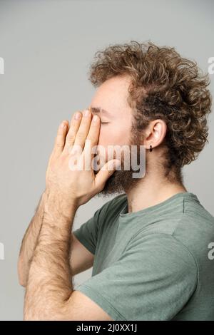 Jeune homme aux cheveux bouclés cache le visage dans les mains fatiguées, concept de migraine. Un gars sympathique ressent de la douleur ou des maux de tête. Jeune homme frustré tryi Banque D'Images