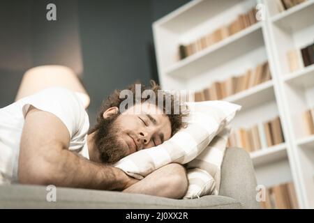 Dormir sur le canapé beau jeune homme barbu embrassant un oreiller après de longues heures de travail tard le soir. Malade jeune homme dormant o Banque D'Images