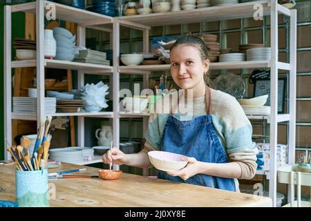 Jeune femme qualifiée en tablier debout à la table et dessin sur un bol en céramique dans un atelier de poterie Banque D'Images