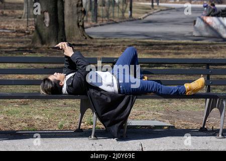 Lors d'une douce journée d'hiver à New York, un homme aux cheveux bicolore lit depuis son téléphone portable tout en étant allongé sur un banc de parc. À Queens, New York. Banque D'Images