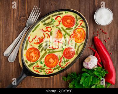 Poêle en fonte ovale avec délicieux chaud omelette aux tomates et poivrons. Brown wood texture background, close-up, vue du dessus. Banque D'Images